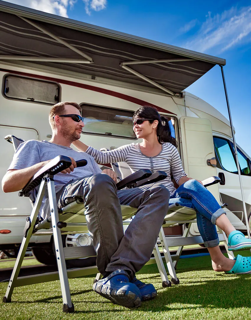 A man and woman sit on outdoor chairs under the awning of a motorhome, smiling at each other on a sunny day.