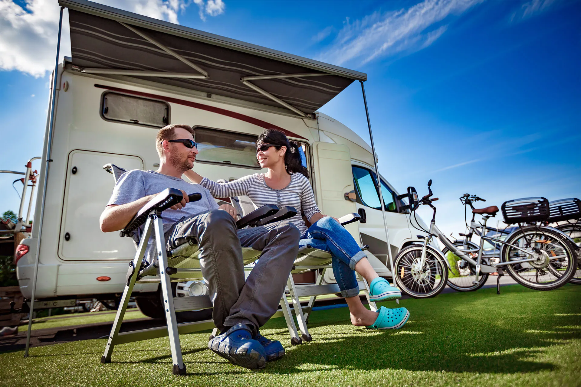 Two people sit on lawn chairs outside a parked RV, with bicycles nearby and an awning providing shade on a sunny day.