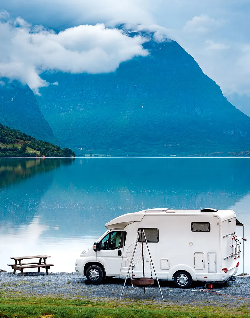 A white camper van parked by a calm lake with a picnic table and swing nearby; large mountains and clouds are reflected in the water in the background.