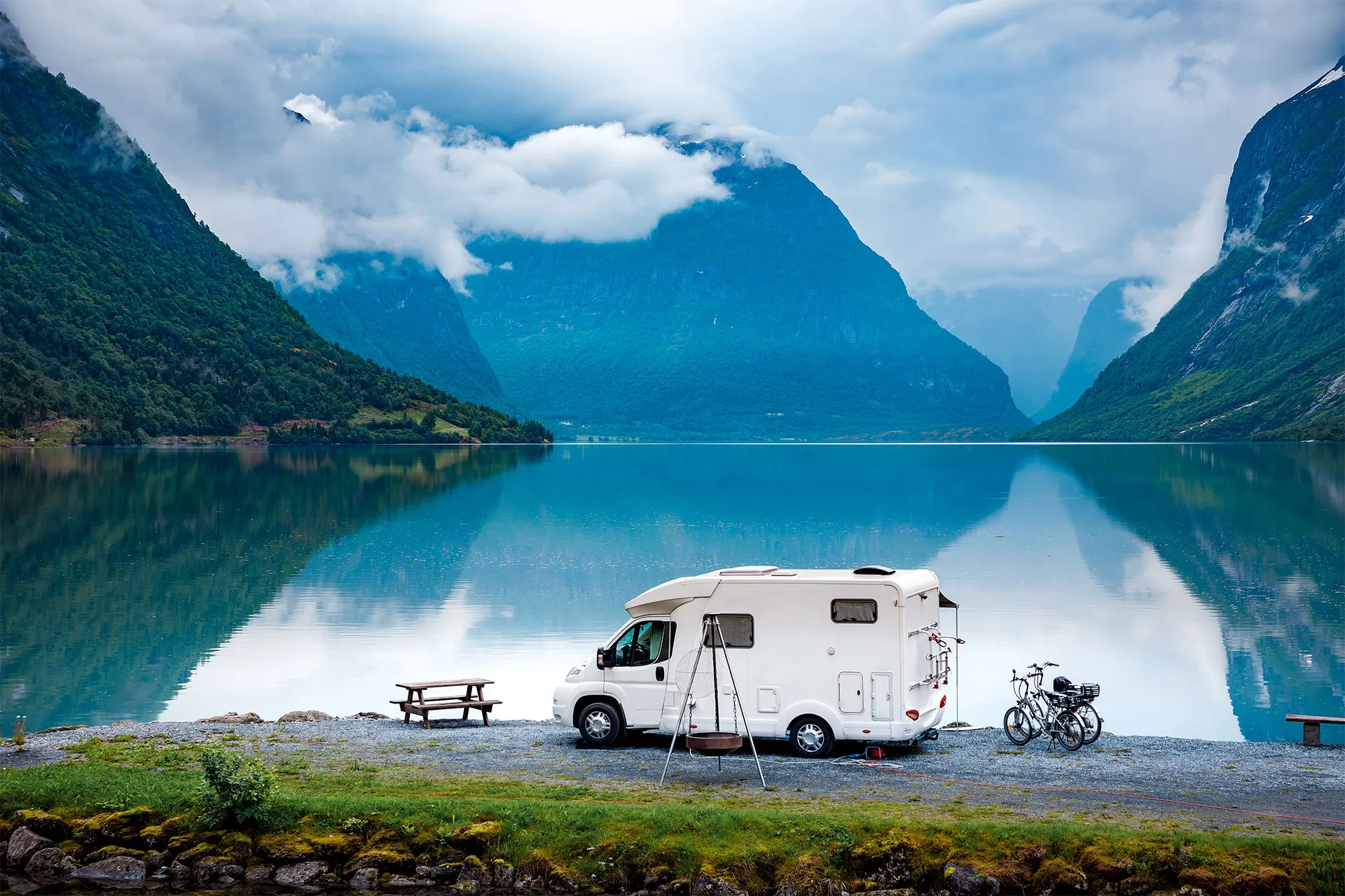 A white camper van is parked by a calm lake with mountains in the background, a picnic table and two bicycles nearby, under a cloudy sky.