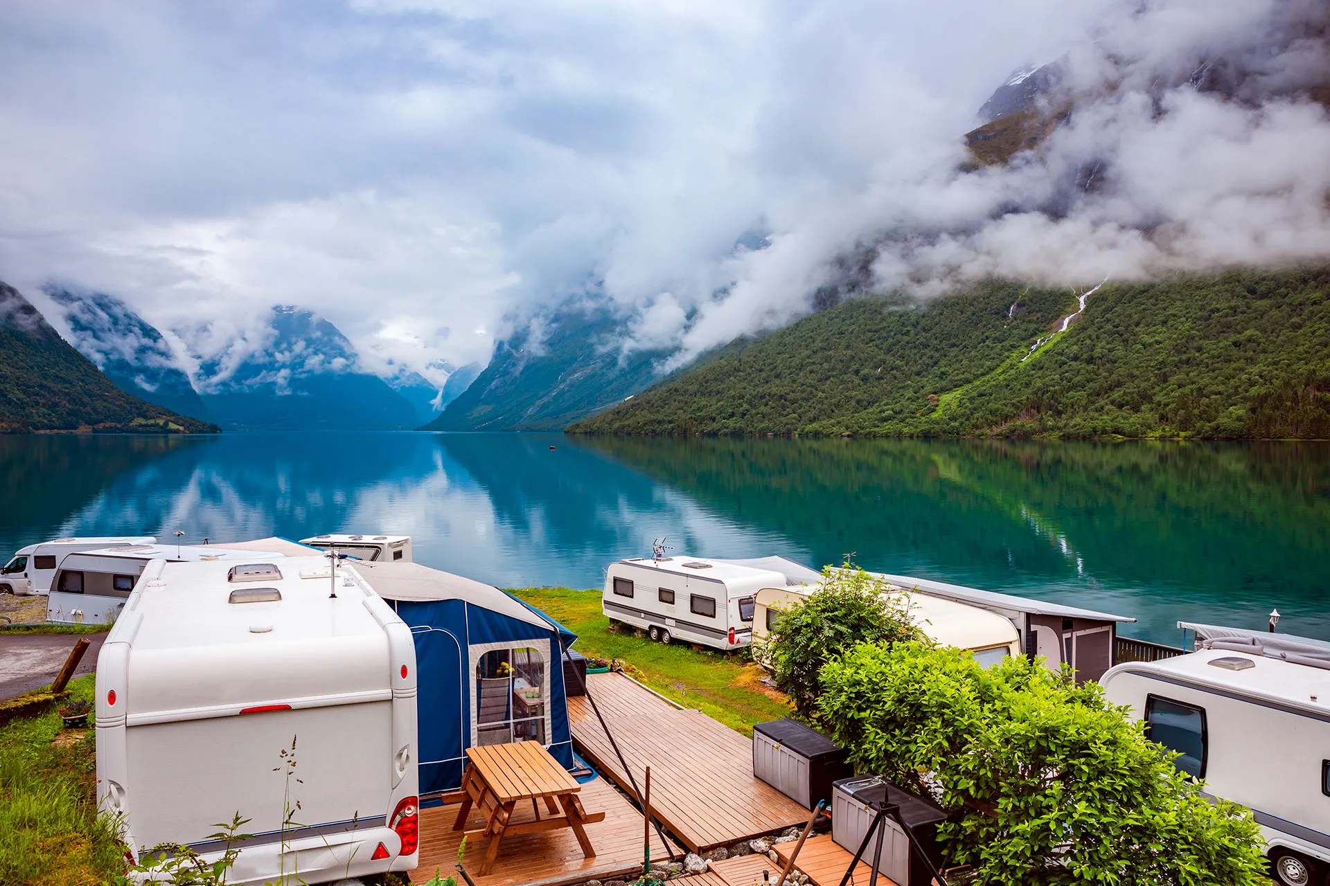 A row of camper vans and tents are parked beside a calm lake surrounded by green mountains with low-hanging clouds.