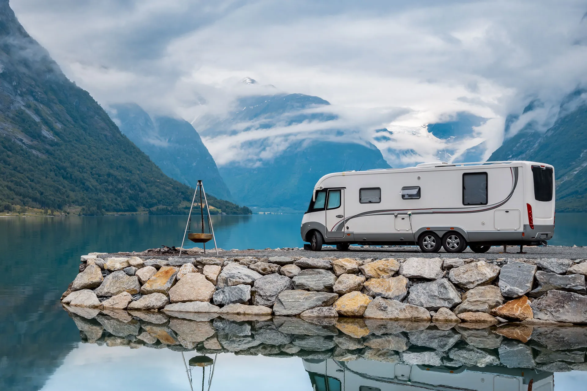 A white camper van is parked next to a lakeshore, with a stone wall, tripod grill, calm water, and mountains with clouds in the background.